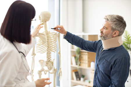 Focus on bearded man wearing C collar placing pen on neck vertebrae while young woman looking at skeleton. Male patient in casual clothes asking physician about cervical spine in doctor's office.